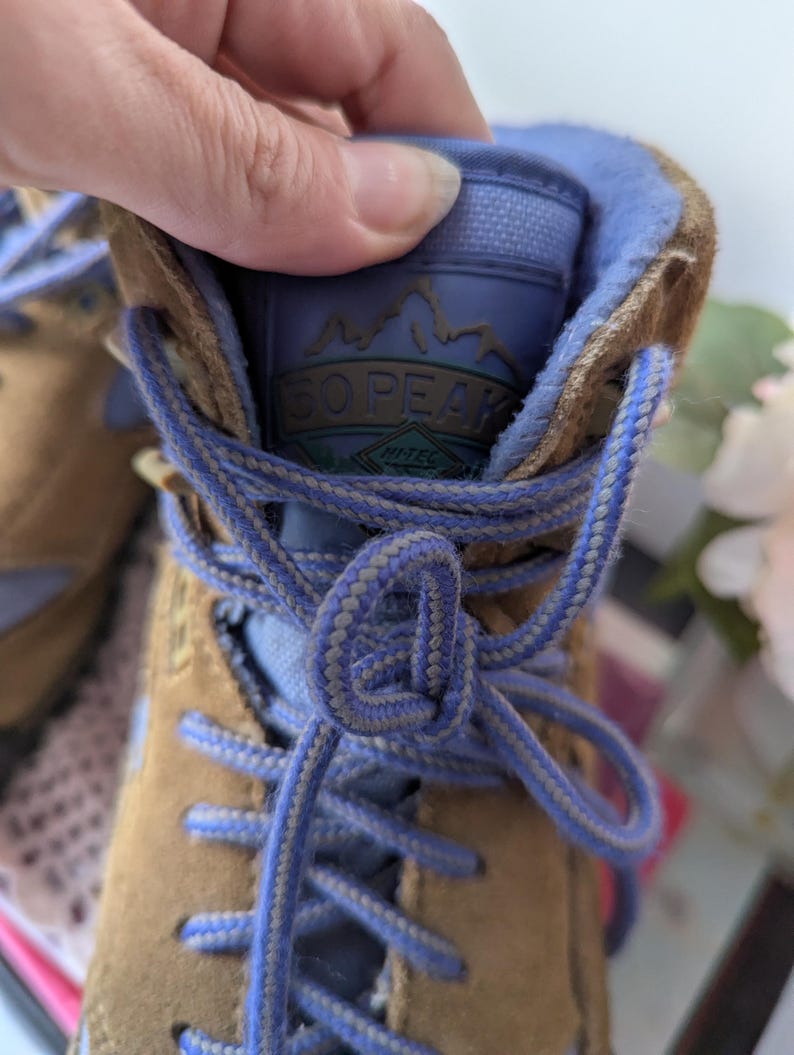 May include: Tan leather hiking boots with blue laces and a blue lining. The boots have a logo on the tongue that says "SO PEAK" with the words "Hi-Tec" underneath.