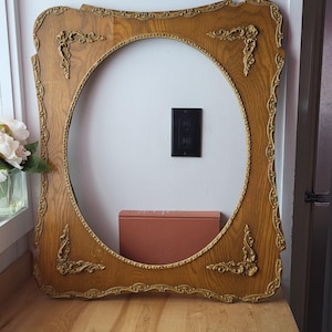 May include: An antique wooden picture frame with an oval aperture. The frame is light brown with gold-coloured decorative details. It rests on a light-coloured wooden surface. The frame's dimensions are approximately 76 cm x 61 cm.