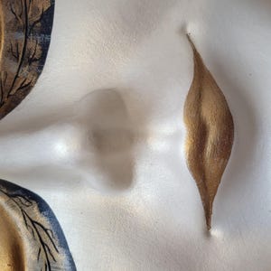 May include: A white ceramic mask with gold and black painted details. The mask features a stylized face with a closed eye, a nose, and a closed mouth. The mouth is painted gold and has a leaf-like shape.
