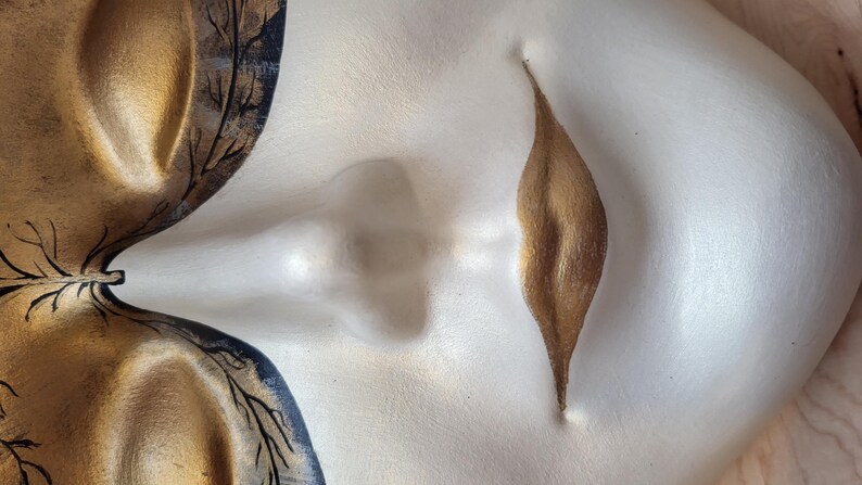 May include: A white ceramic mask with gold and black painted details. The mask features a stylized face with a closed eye, a nose, and a closed mouth. The mouth is painted gold and has a leaf-like shape.