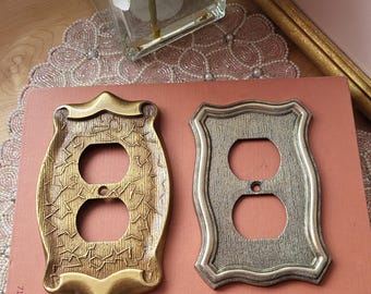 May include: Two decorative electrical outlet covers. One is gold-toned with a textured pattern, the other is silver-toned with a similar texture. Both have a shield-like shape and two circular openings for the outlets.