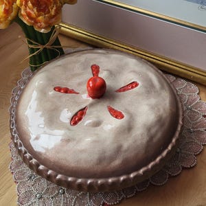 Puede incluir: Una fuente de cerámica para pastel con un esmalte marrón claro y una corteza decorativa. El pastel tiene un diseño de cereza en la parte superior, con una cereza roja en el centro y detalles rojos. El plato está sobre un mantel individual con cuentas.