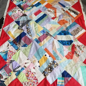 Op de afbeelding: Een kleurrijk patchwork-deken met een rode rand en witte franjes. Het deken is gemaakt van verschillende stoffen in verschillende kleuren en patronen, waaronder bloemen, geometrische vormen en ruitjes.