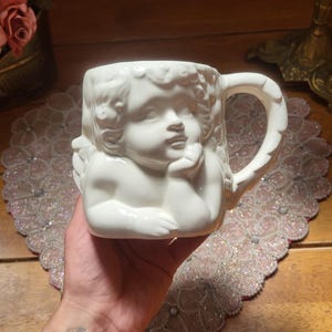 May include: White ceramic mug featuring a sculpted cherub design. The mug has a handle and is held in a hand. The cherub's face is detailed, with curly hair and a thoughtful expression. The mug is a decorative item.