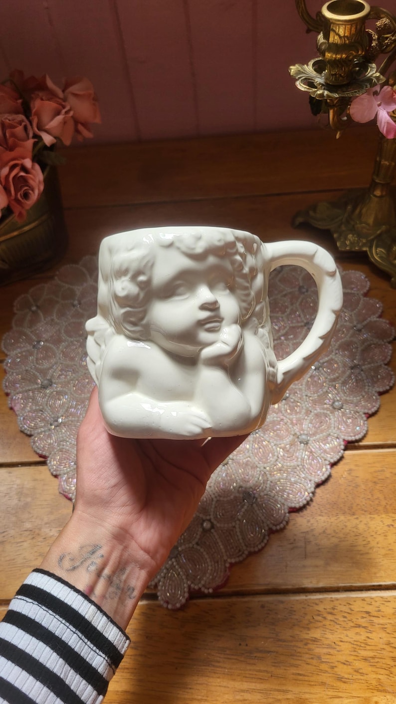 May include: White ceramic mug featuring a sculpted cherub design. The mug has a handle and is held in a hand. The cherub's face is detailed, with curly hair and a thoughtful expression. The mug is a decorative item.