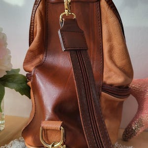 May include: A brown leather satchel bag with a gold-toned handle and a long adjustable strap. The bag has two zippered compartments and a flap closure.