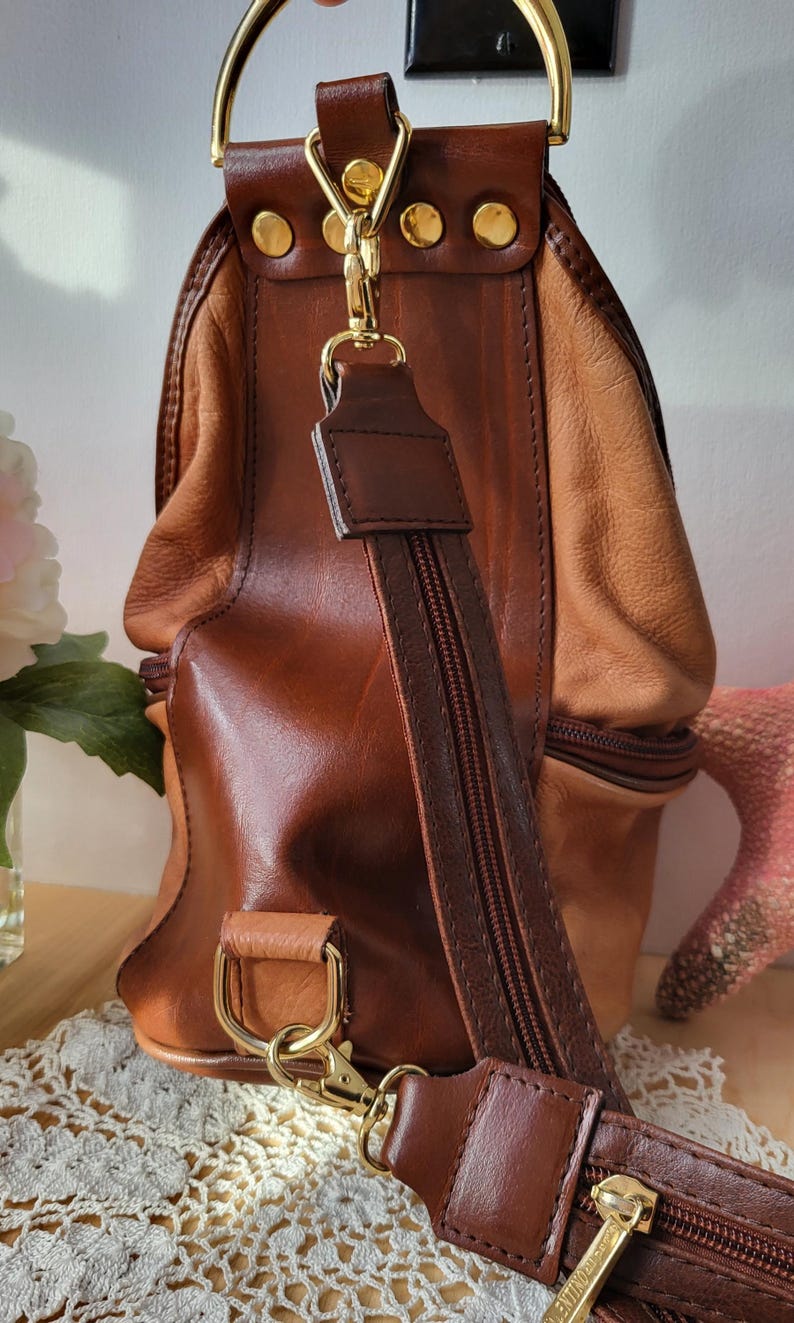 May include: A brown leather satchel bag with a gold-toned handle and a long adjustable strap. The bag has two zippered compartments and a flap closure.