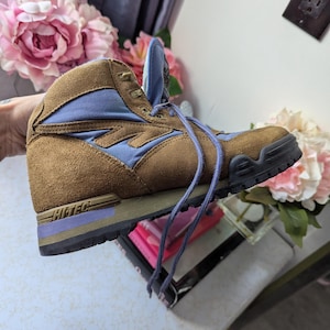 May include: Brown suede hiking boots with blue accents and a purple Hi-Tec logo on the side. The boots have a lace-up closure and a rugged outsole.