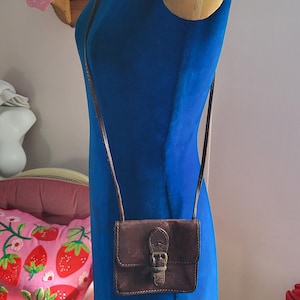 Op de afbeelding: Een kleine, bruine leren crossbodytas met een gespsluiting wordt getoond op een blauwe mannequin. De tas heeft een lange, dunne riem. De mannequin staat voor een roze muur met decoratieve items.