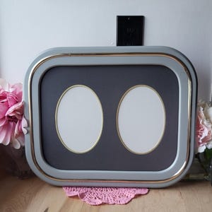 May include: A grey double picture frame with gold trim and two oval openings. The frame has a dark grey mount and sits on a wooden surface. A pink doily is beneath the frame.