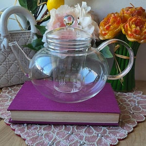 Puede incluir: Una tetera de vidrio transparente con cuerpo redondo y tapa de vidrio, apoyada sobre un libro morado. La tetera tiene un infusor incorporado. El fondo incluye limones, flores y un bolso decorativo.
