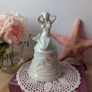 Può includere: Una campana in ceramica bianca iridescente con una statuetta di sirena in cima. La sirena è seduta su un delfino e ha le mani sulla testa. La campana è decorata con delfini e conchiglie.