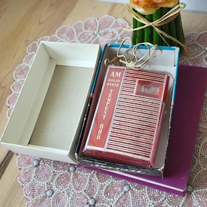 May include: A vintage red and white AM Tempest 888 solid state radio in its original box. The radio is in a black box with blue trim, and the text "TEMPEST 888" is visible. The radio is on top of a purple book.