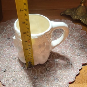 May include: A white ceramic mug with a textured exterior, featuring a handle. The mug is approximately 4 inches (10 cm) tall. It sits on a beaded placemat with a pink and white floral design. A yellow measuring tape is next to the mug.