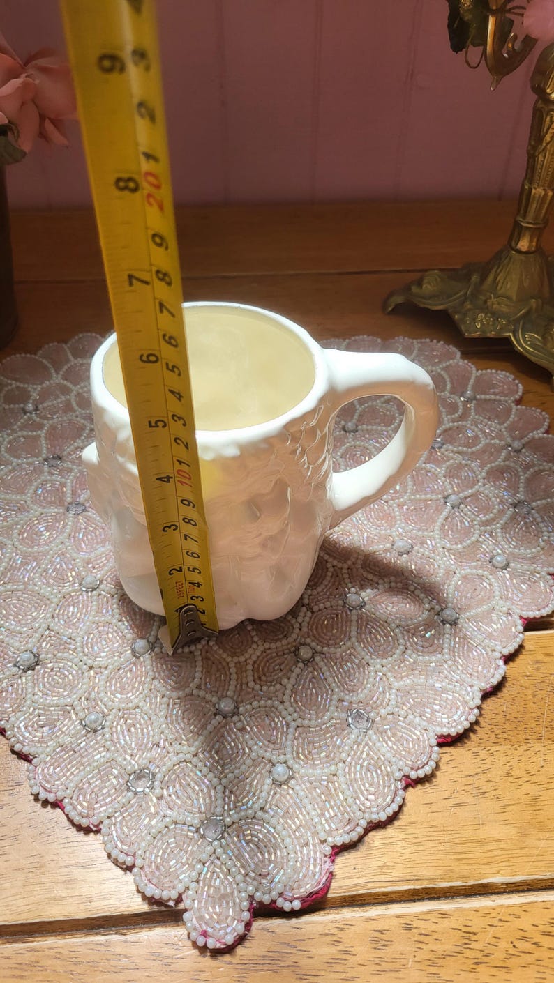 May include: A white ceramic mug with a textured exterior, featuring a handle. The mug is approximately 4 inches (10 cm) tall. It sits on a beaded placemat with a pink and white floral design. A yellow measuring tape is next to the mug.