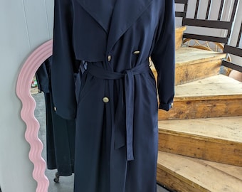 May include: A navy blue trench coat with a double-breasted front, a matching belt, and gold-colored buttons. The coat is displayed on a mannequin. The coat has a classic design and is made of a smooth fabric.