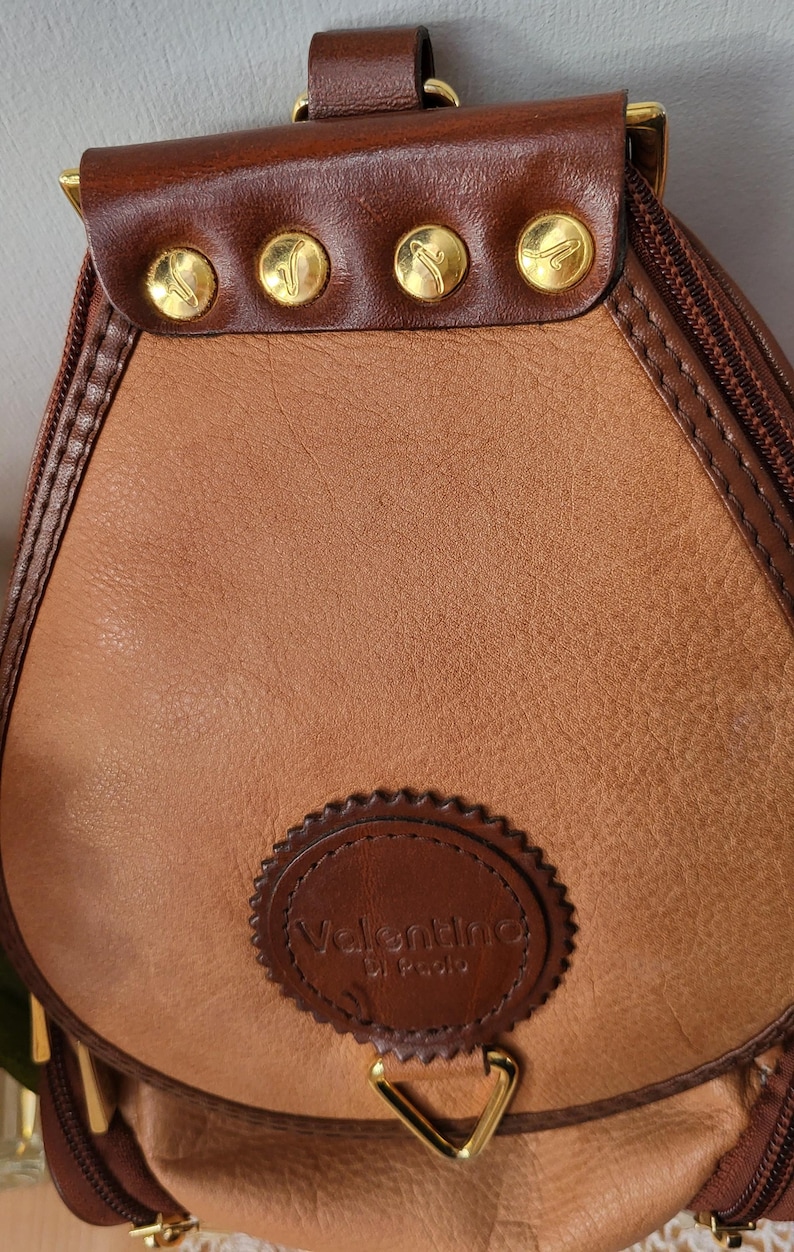 May include: A brown leather satchel bag with gold accents and a circular logo that reads "Valentino di Paolo".