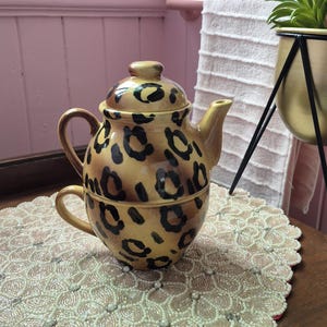 May include: A ceramic tea for one set featuring a teapot with a lid and a matching teacup stacked together. The set has a glossy tan finish with a black leopard print pattern throughout. The teapot has a curved handle and a spout, while the teacup has a handle.