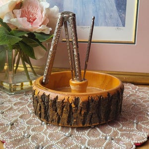 Pode incluir: Um conjunto de quebra-nozes de madeira com borda de casca natural. O conjunto inclui um quebra-nozes prateado e duas palhetas, todos em uma tigela redonda de madeira. O fundo apresenta uma impressão emoldurada e um vaso de flores cor-de-rosa.