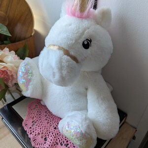 May include: White plush unicorn toy with pink mane and gold horn. The unicorn has sparkly iridescent sequins on its hooves and a gold ribbon around its muzzle.