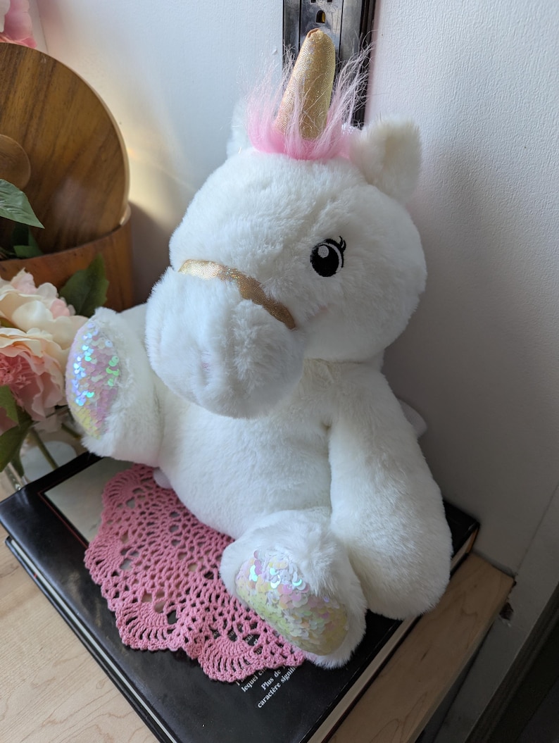 May include: White plush unicorn toy with pink mane and gold horn. The unicorn has sparkly iridescent sequins on its hooves and a gold ribbon around its muzzle.