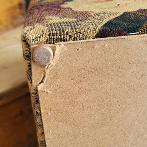 May include: Close-up of the bottom of a fabric-covered storage ottoman. The ottoman is made of wood and has a fabric top with a floral pattern. The fabric is slightly torn at the corner, revealing the wood underneath. A metal nail is visible on the side of the ottoman.