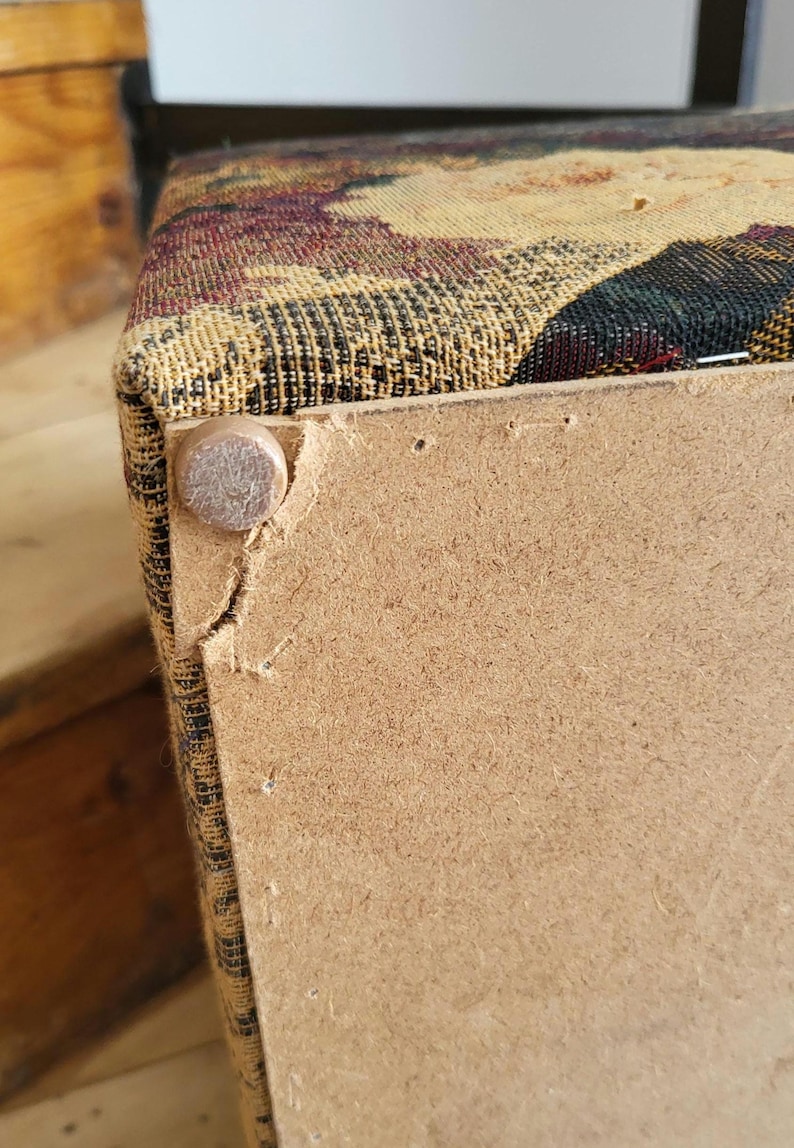 May include: Close-up of the bottom of a fabric-covered storage ottoman. The ottoman is made of wood and has a fabric top with a floral pattern. The fabric is slightly torn at the corner, revealing the wood underneath. A metal nail is visible on the side of the ottoman.