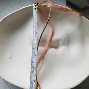 May include: A white ceramic mask with a gold and pink ribbon attached to the back. The mask is 8 inches in diameter.