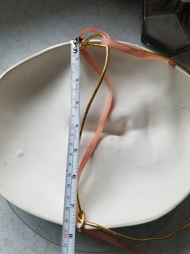 May include: A white ceramic mask with a gold and pink ribbon attached to the back. The mask is 8 inches in diameter.