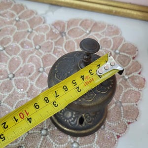 Puede incluir: Campana de lat&oacute;n antiguo con detalles florales, de aproximadamente 10 cm de di&aacute;metro. La campana tiene un peque&ntilde;o asa en la parte superior y se mide con una cinta m&eacute;trica amarilla. La campana est&aacute; sobre un mantel individual rosa con cuentas.