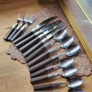 May include: A set of stainless steel silverware with brown handles. The set includes forks, knives, and spoons. The silverware is arranged on a wooden surface, with a decorative doily and framed artwork in the background.