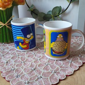May include: Two white ceramic mugs featuring cartoon characters. One mug has a blue background with Bart Simpson, and the other has Homer Simpson holding a burger. The mugs are on a pink beaded placemat.