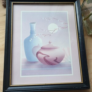 Op de afbeelding: Een ingelijste print van een stilleven met een blauwe vaas en een roze pot met deksel met een bloemenmotief. De achtergrond is zacht paars met een witte maan en een tak met witte bloesems.