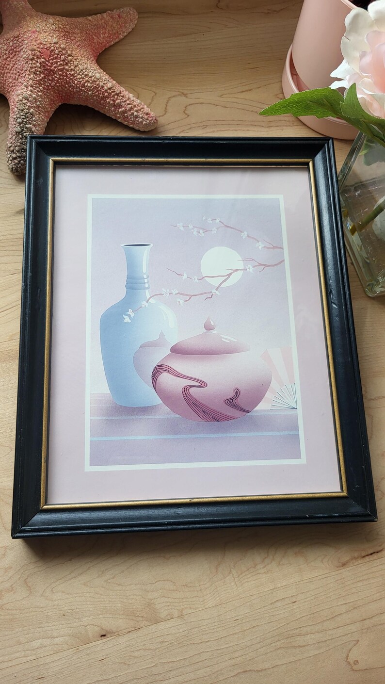 Op de afbeelding: Een ingelijste print van een stilleven met een blauwe vaas en een roze pot met deksel met een bloemenmotief. De achtergrond is zacht paars met een witte maan en een tak met witte bloesems.