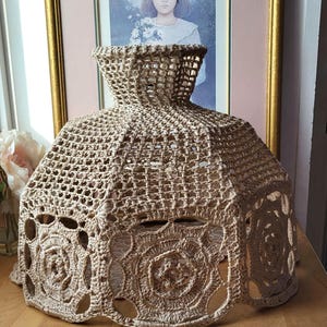 May include: A beige, crocheted lampshade with a flared, bell-like shape. The lampshade features an open weave design with decorative circular cutouts near the base. The lampshade is sitting on a wooden surface.
