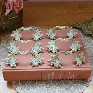 May include: Six vintage-style drawer pulls with a white and gold finish, arranged on a pink book. Each pull features a curved handle with ornate leaf detailing. The pulls are likely made of metal and are suitable for furniture restoration projects.