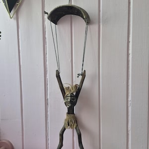 Könnte beinhalten: Eine dunkelbraune Holzfigur einer Person mit erhobenen Armen, die an dünnen dunklen Schnüren von einem geschwungenen Holzdach hängt. Die Figur hat ein geschnitztes Gesicht und einen hellbraunen Faserrock um die Taille und hängt vertikal.