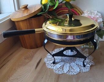 May include: A gold chafing dish with a black handle and a black metal stand. The chafing dish is on a white crocheted doily.