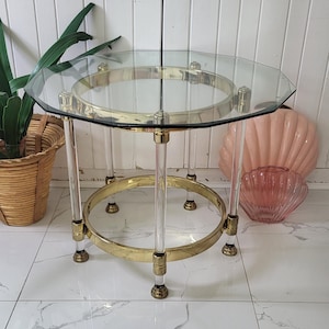 Puede incluir: Una mesa auxiliar de cristal transparente y latón. La mesa tiene una tapa de cristal octogonal transparente, soportada por patas de acrílico transparente y anillos de latón. La mesa tiene aproximadamente 61 cm de diámetro y 51 cm de altura.