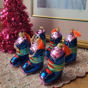 May include: Six vintage Christmas boot ornaments in blue and pink with orange tassels. The boots have gold, green, and red detailing. A pink tinsel tree and a framed print are in the background.