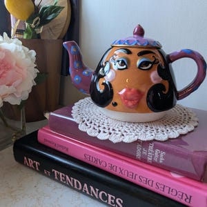 May include: A ceramic teapot with a painted face, black hair, and purple and blue accents. The teapot is sitting on a stack of books with French titles.