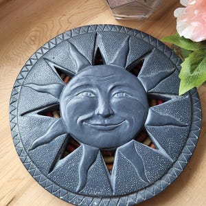 Może przedstawiać: Niebieska ceramiczna słońce z uśmiechającą się twarzą i szczegółowym wzorem. Słońce jest otoczone pierścieniem mniejszych słońc, z których każde ma ostry promień.