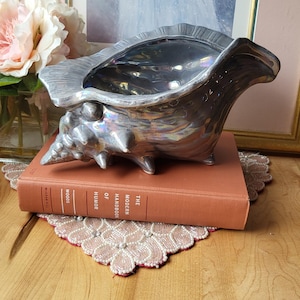Puede incluir: Un plato decorativo plateado en forma de concha. El plato está sobre un libro antiguo titulado "The Modern Handbook of Humor". El fondo incluye una pintura enmarcada y flores rosas.
