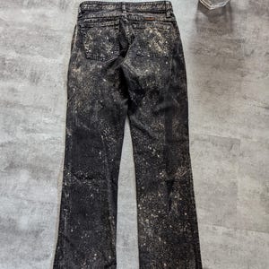 May include: Black denim jeans with a bleach splatter pattern. The jeans have a bootcut leg style.