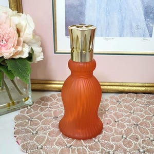 May include: An orange glass fragrance lamp with a gold-colored metal top. The lamp has a ribbed design and a flared base. It is sitting on a beaded floral placemat, with a framed painting in the background.