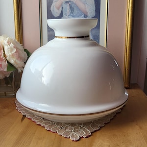 May include: A white, dome-shaped lamp shade with a gold trim. The lamp shade sits on a decorative, beaded doily. A painting of a girl holding flowers is in the background.