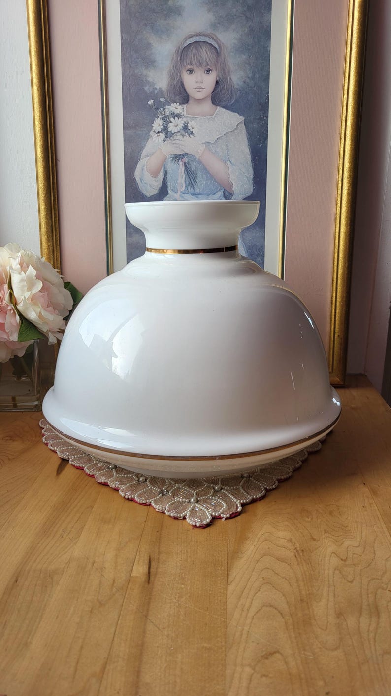 May include: A white, dome-shaped lamp shade with a gold trim. The lamp shade sits on a decorative, beaded doily. A painting of a girl holding flowers is in the background.