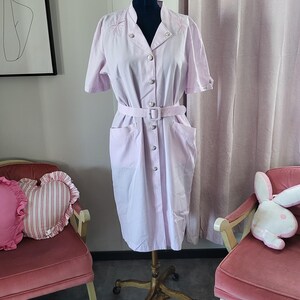 May include: A light pink dress with short sleeves and a button-up front. The dress has a belt and two pockets. The dress is on a mannequin and is displayed against a white wall.