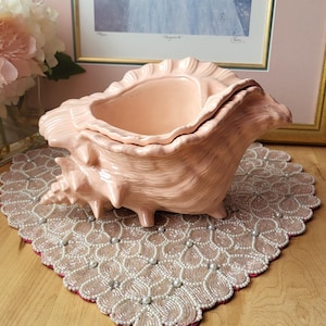 Puede incluir: Un recipiente de cerámica rosa claro en forma de concha con tapa, sobre un mantel individual con cuentas. El recipiente tiene una superficie texturizada y está sobre un fondo rosa con una imagen enmarcada.