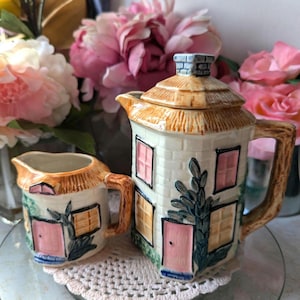 May include: A ceramic teapot and creamer set shaped like houses with brown roofs, pink doors, and green window trim. The teapot has a chimney and the creamer has a small window. The set is on a white doily.